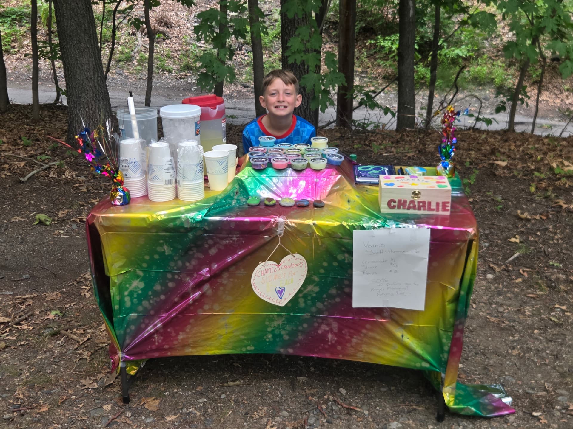 Charlie sitting behind a rainbow-covered table selling lemonade, slimes, and painted rocks on the bike path