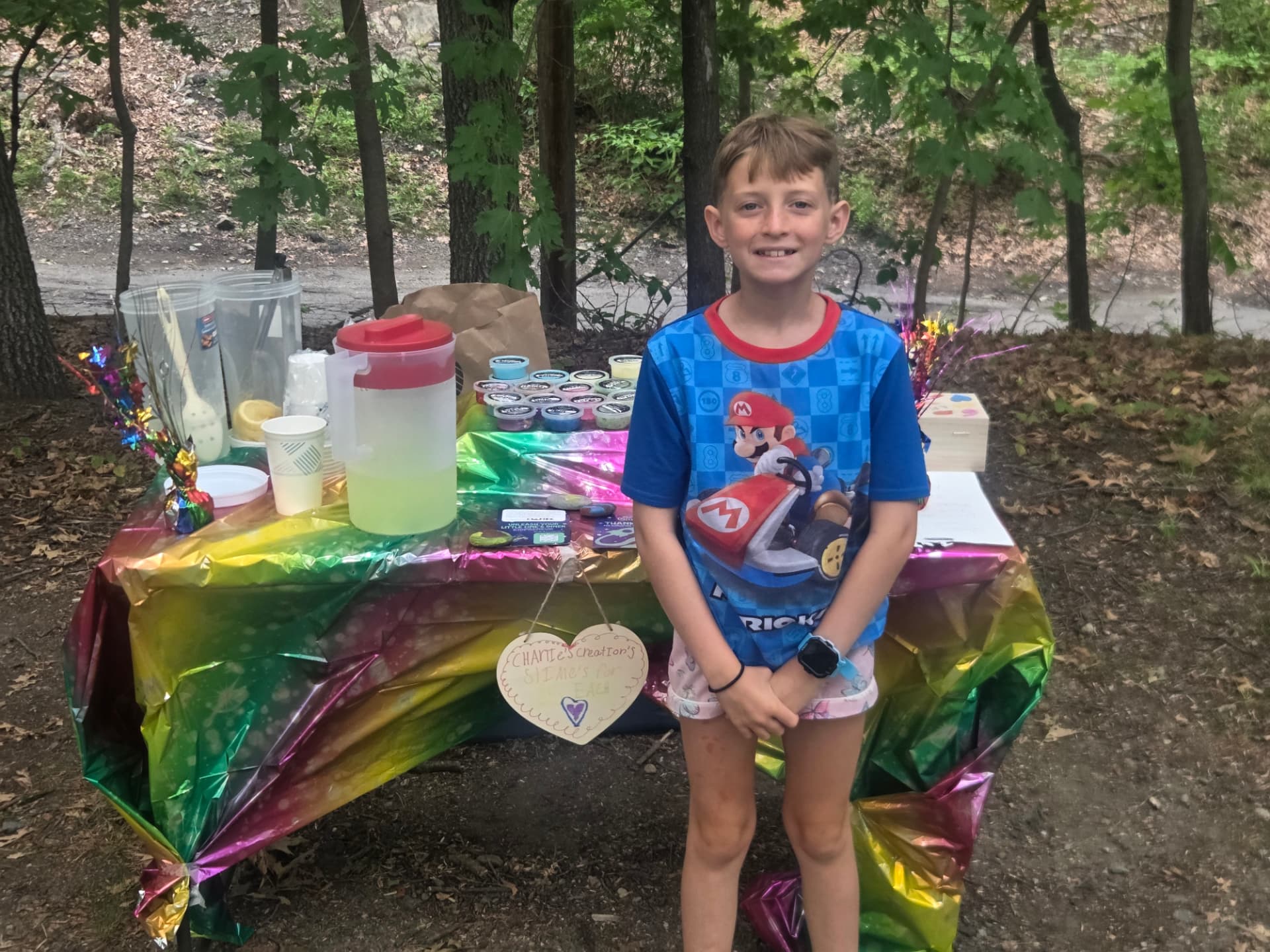 Charlie standing proudly next to her lemonade and slime stand in the woods